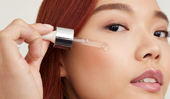 Close-up of a woman applying a serum to her cheek using a dropper. The woman has smooth skin, light makeup, and a subtle smile. 