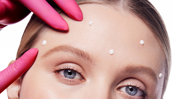 A pink medical gloved hand pointing to Dynamic Wrinkle Limiter topical cream on a blonde-haired woman’s upper face