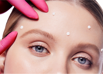 A pink medical gloved hand pointing to Dynamic Wrinkle Limiter topical cream on a blonde-haired woman’s upper face